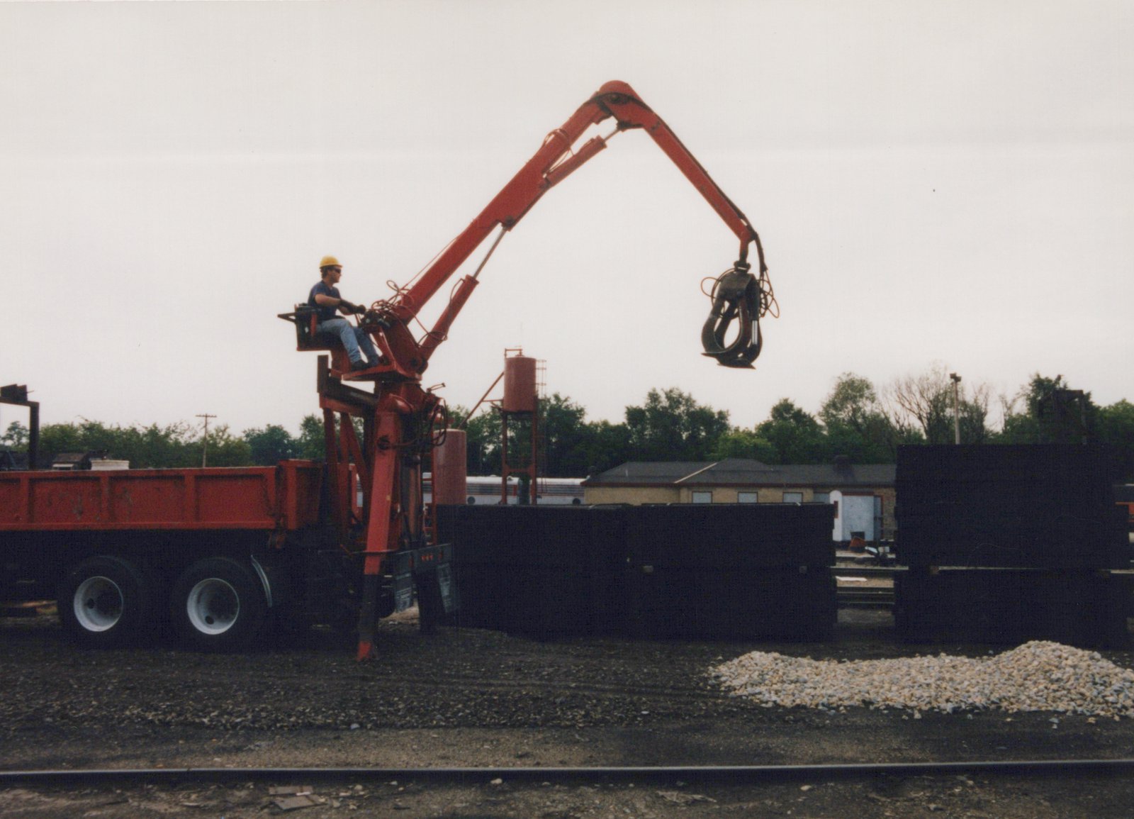 A red truck with a tall red hydraulic crane arm and a dangling black grapple stands next to dark stacks of railroad ties; a worker sits at the crane controls, and a red water tank, a low brick building and a silver passenger car appear in the background under a gray sky.