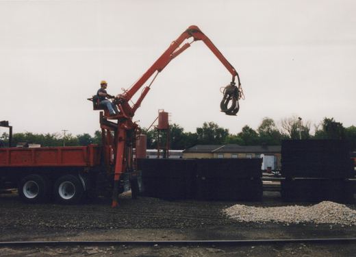 A red truck with a tall red hydraulic crane arm and a dangling black grapple stands next to dark stacks of railroad ties; a worker sits at the crane controls, and a red water tank, a low brick building and a silver passenger car appear in the background under a gray sky.