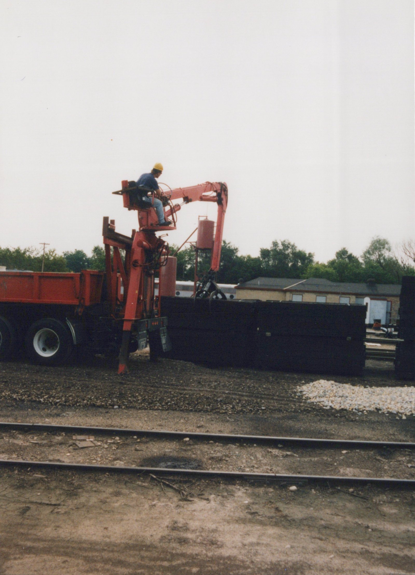 A side view of a railroad yard on an overcast day, showing a red mobile crane with an operator in a yellow hard hat, a black freight car, and two parallel tracks in the foreground.