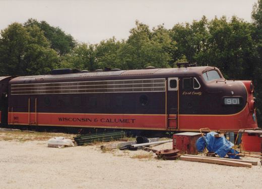 Side view of a maroon and red-orange cab-unit diesel locomotive, number 901, lettered Wisconsin & Calumet, with tarps, lumber, pipes and an old red cabinet on the gravel in front of it and trees behind.