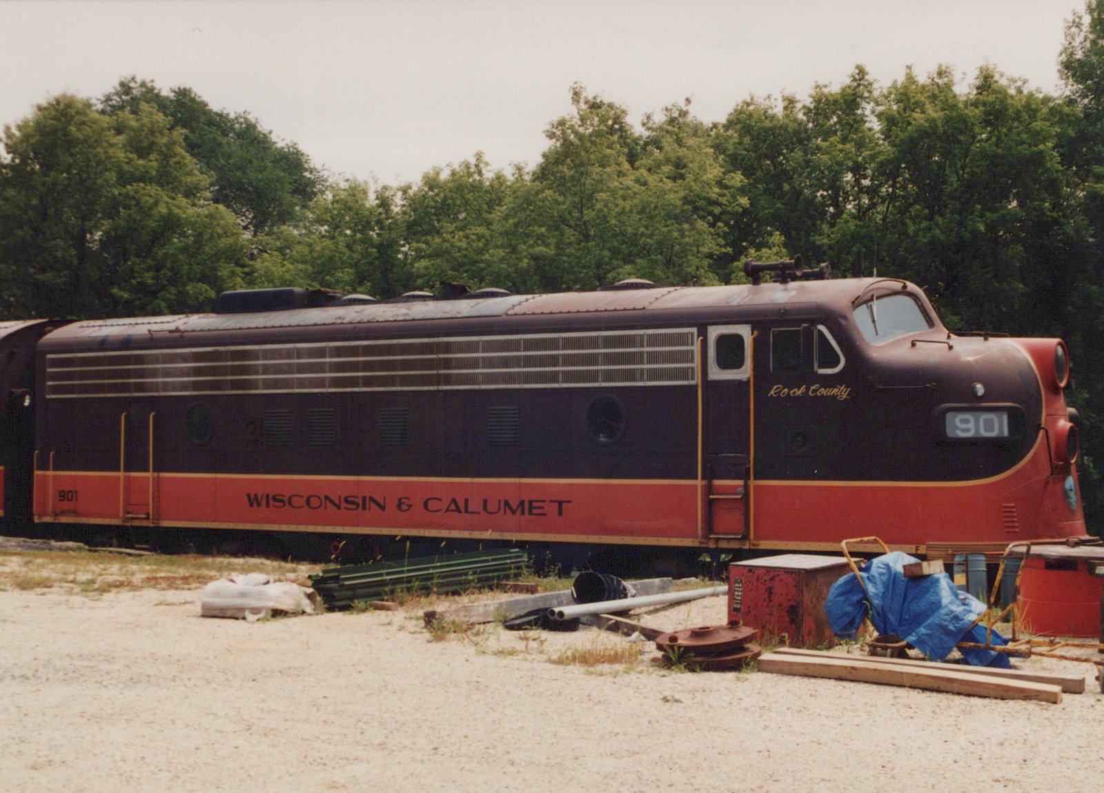 Side view of a maroon and red-orange cab-unit diesel locomotive, number 901, lettered Wisconsin & Calumet, with tarps, lumber, pipes and an old red cabinet on the gravel in front of it and trees behind.
