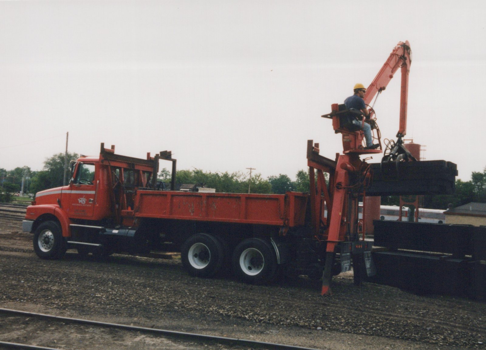 Side view of a red tandem-axle dump truck with a rear-mounted knuckle-boom crane on gravel beside a track. A worker in a hard hat sits in the crane seat and lifts a stack of dark ties. Trees, a red tank and part of a silver passenger car are in the background.