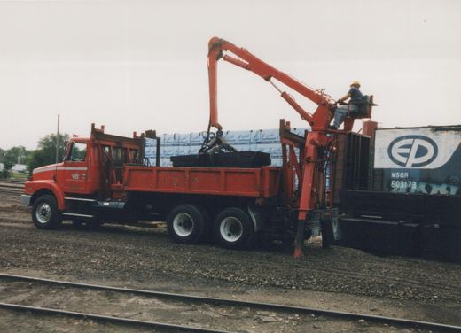 A red tandem-axle truck with a large red knuckle-boom crane stands on gravel beside yard tracks. A worker in a yellow hard hat sits at the crane controls, and its grapple rests on a load of black crossties. A blue boxcar with an EP herald is at the right.