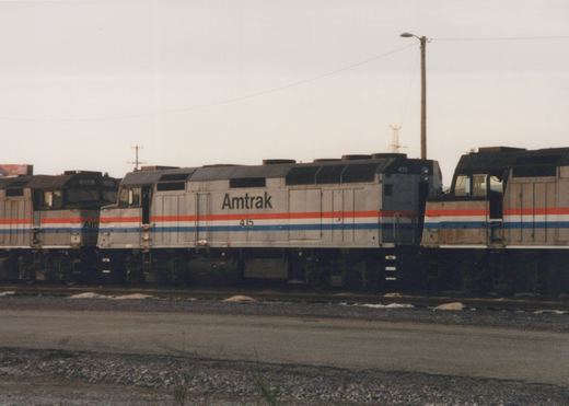 A silver diesel locomotive lettered Amtrak with the number 415 and red, white and blue stripes is coupled between two similar locomotives on a gravel-edged yard track under a grey sky.