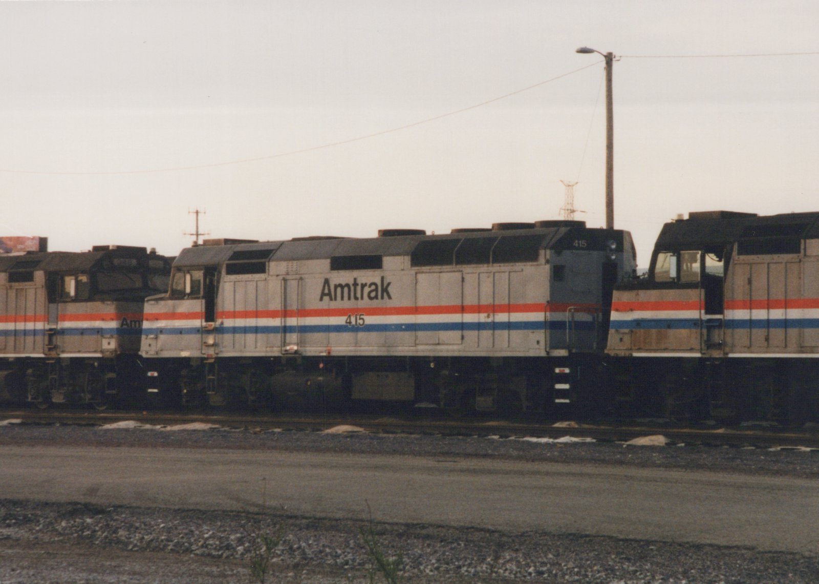 A silver diesel locomotive lettered Amtrak with the number 415 and red, white and blue stripes is coupled between two similar locomotives on a gravel-edged yard track under a grey sky.
