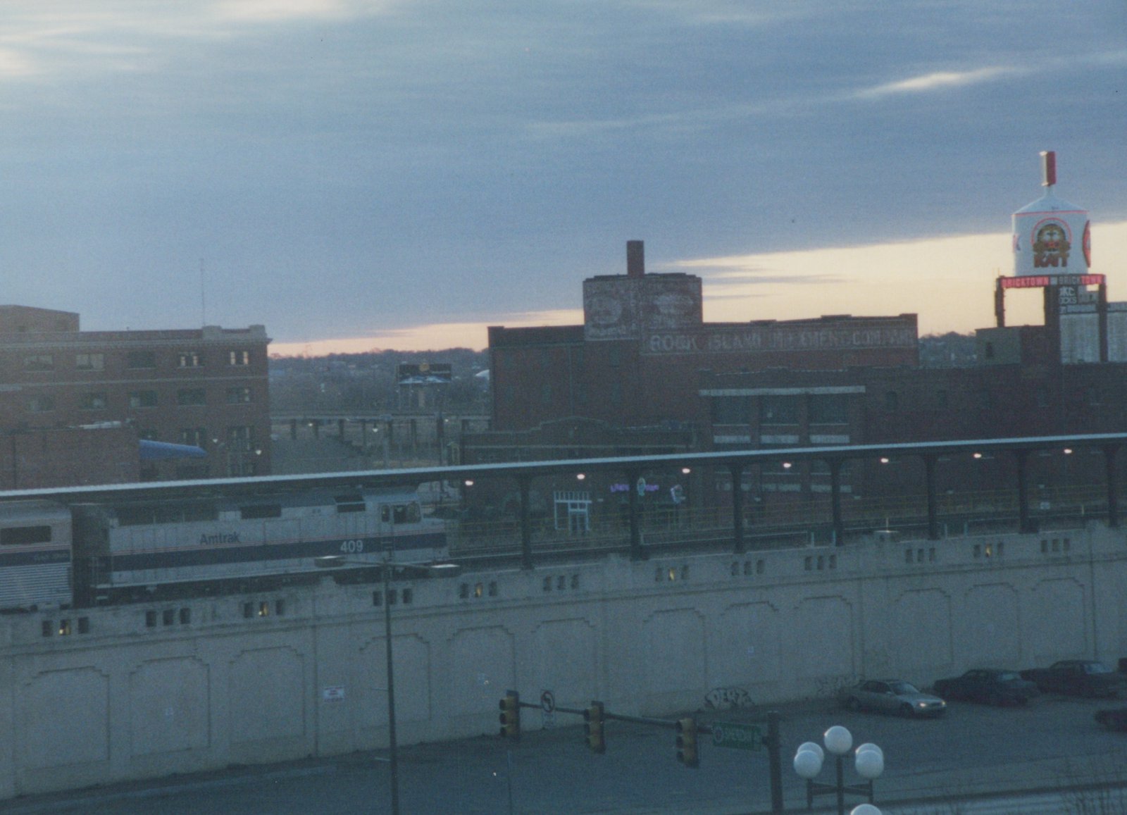A silver Amtrak diesel locomotive numbered 409 and a passenger car stand at a long canopied platform on a concrete wall, with brick warehouses, a lit water tower and a cloudy sky behind.