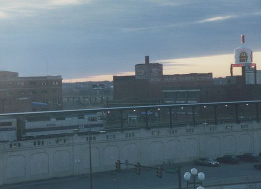A silver Amtrak diesel locomotive numbered 409 and a passenger car stand at a long canopied platform on a concrete wall, with brick warehouses, a lit water tower and a cloudy sky behind.