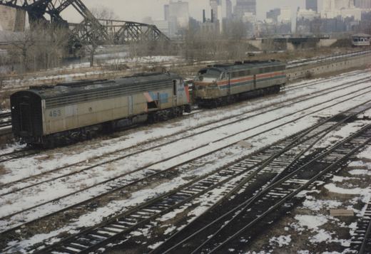 Two weathered silver diesel locomotives with Amtrak stripes stand on a snow-covered yard track. Behind them are a large steel truss bridge, bare trees and city skyscrapers.