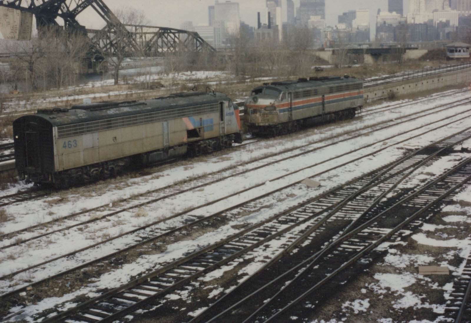 Two weathered silver diesel locomotives with Amtrak stripes stand on a snow-covered yard track. Behind them are a large steel truss bridge, bare trees and city skyscrapers.