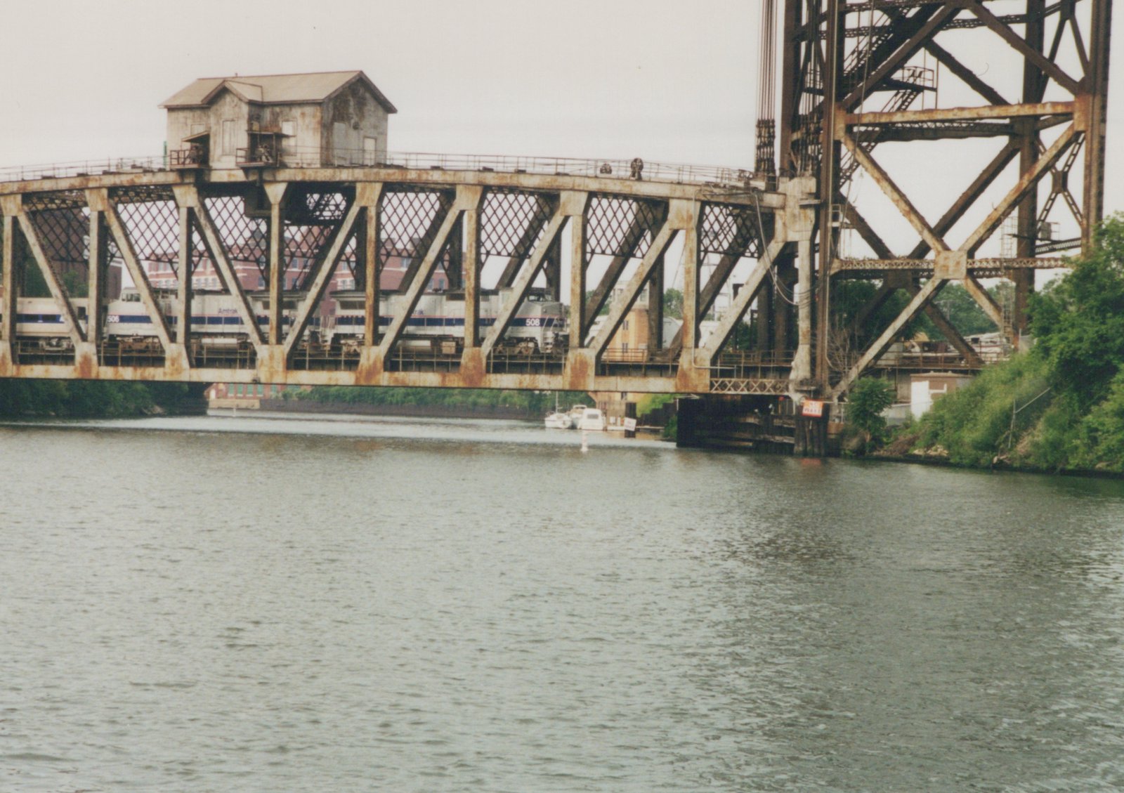 Two silver Amtrak diesel locomotives with blue stripes seen through the lattice truss of a rusty swing bridge with a small tender's house on top, beside a tall lift bridge tower, with the river in the foreground.