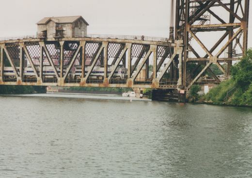 Two silver Amtrak diesel locomotives with blue stripes seen through the lattice truss of a rusty swing bridge with a small tender's house on top, beside a tall lift bridge tower, with the river in the foreground.