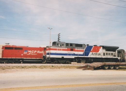 A red, white and blue Amtrak diesel locomotive numbered 513 coupled behind a red CP Rail System locomotive, with a silver passenger car behind it and a flatbed trailer parked beside the track.