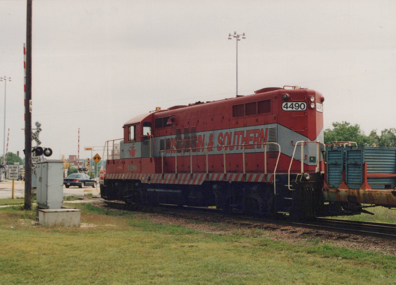 A red and gray diesel locomotive lettered Wisconsin & Southern, numbered 4490, coupled to a rusty blue freight car beside a road crossing with gates and signals under an overcast sky.