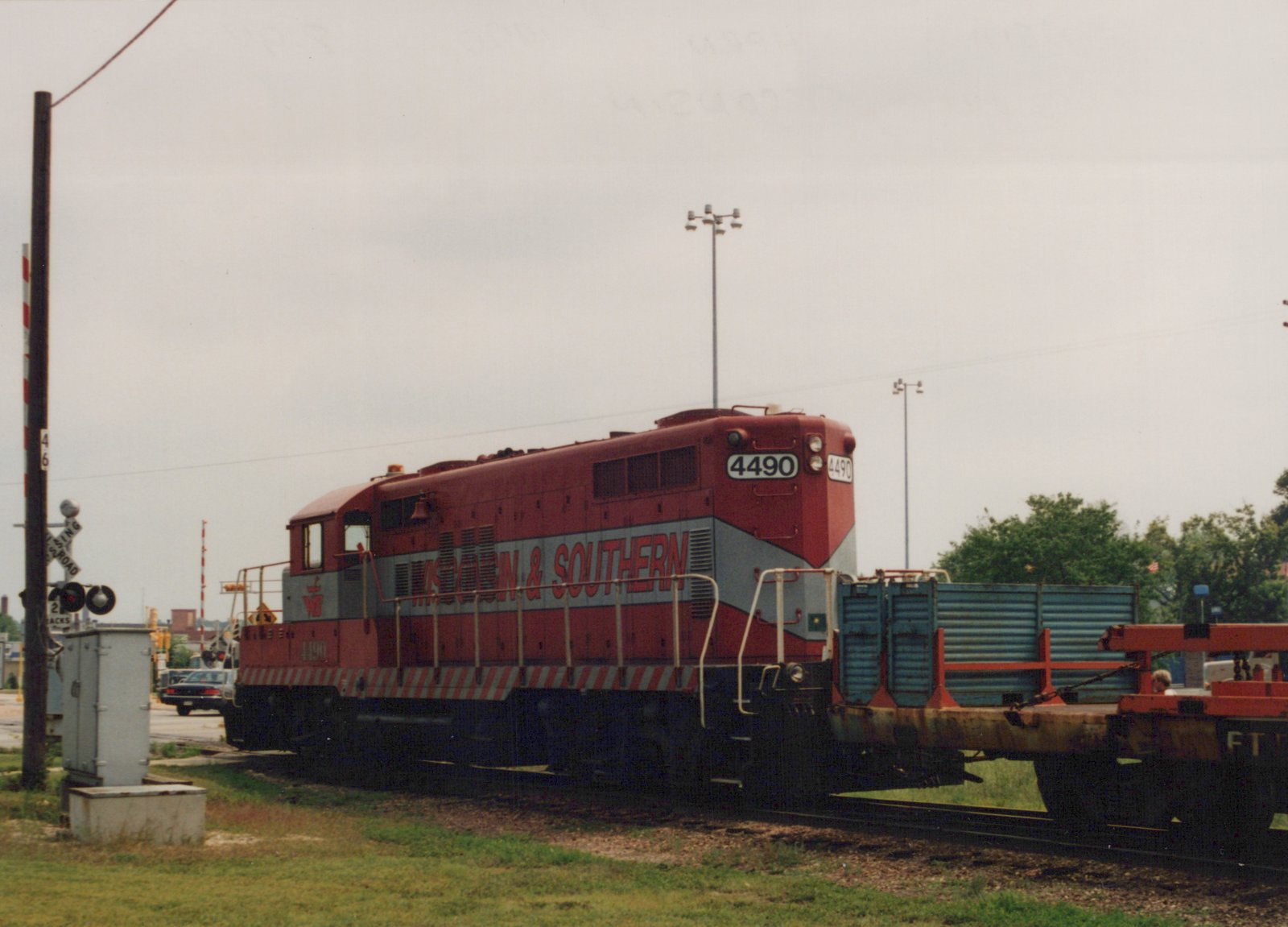 A red and grey diesel locomotive numbered 4490, coupled to a blue bulkhead flatcar and a red-orange flatcar, beside a railroad crossing with signals and a waiting car.