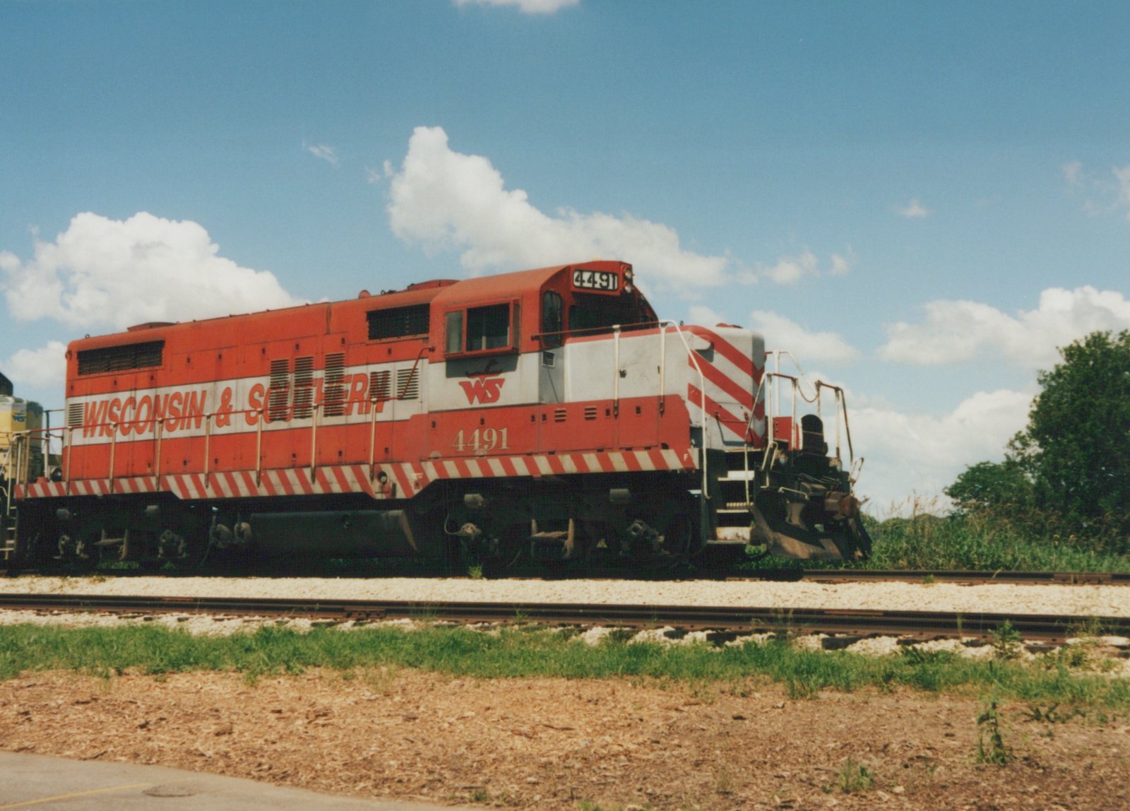 A red-orange and white diesel locomotive numbered 4491, lettered Wisconsin & Southern, stands on a track beside a second track under a blue sky with clouds, with a yellow unit coupled behind it.