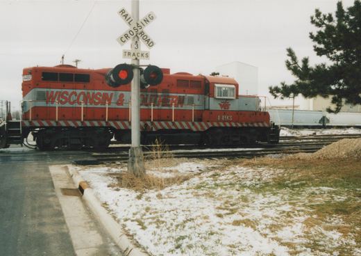 A red and gray Wisconsin & Southern diesel locomotive numbered 4493 seen from the side behind a railroad crossing signal with one red light lit, with light snow on the grass in the foreground and gray covered hoppers behind.