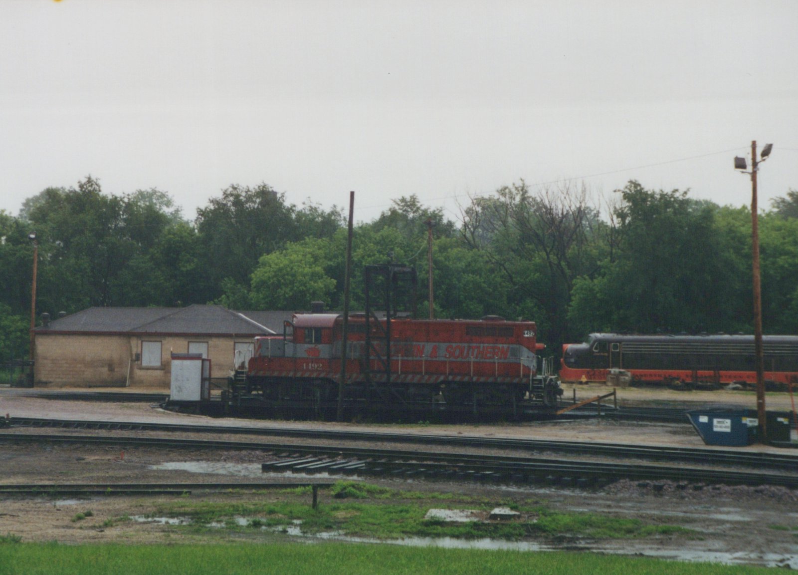 A red diesel locomotive with a silver stripe reading '& SOUTHERN' sits on a turntable in a rain-soaked yard in front of a tan brick building and trees, with a black and orange streamlined locomotive beyond it.