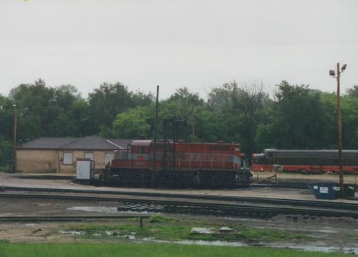 A red diesel locomotive with a silver stripe reading '& SOUTHERN' sits on a turntable in a rain-soaked yard in front of a tan brick building and trees, with a black and orange streamlined locomotive beyond it.
