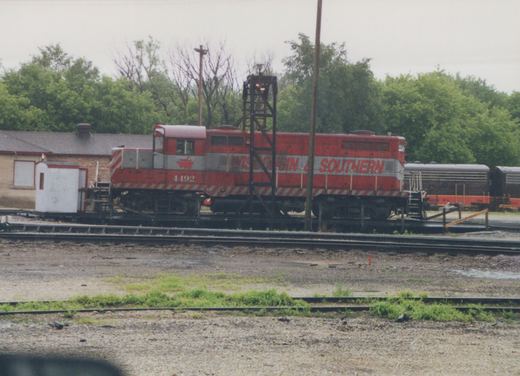 A red and gray diesel locomotive lettered Wisconsin & Southern and numbered 4492 stands on a turntable beside a white shanty and a steel light tower, with two dark locomotives with orange stripes and green trees behind.