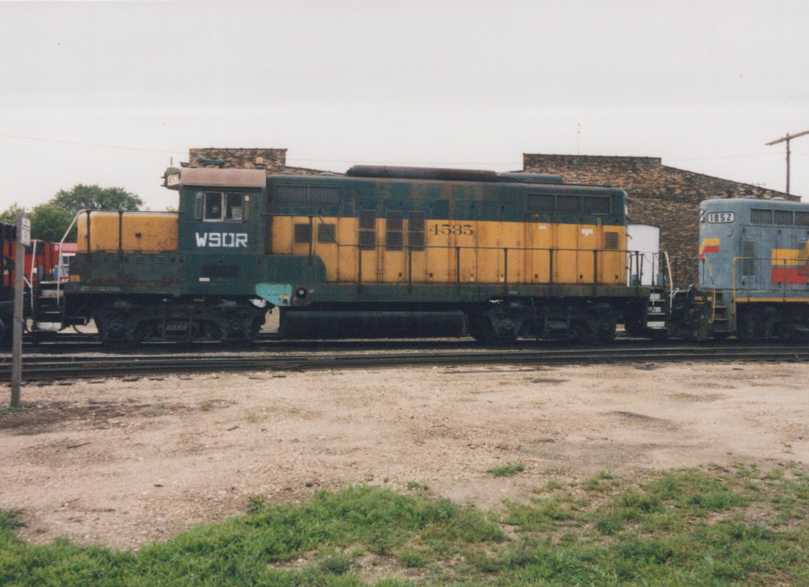 Side view of a weathered yellow and green diesel locomotive lettered WSOR and numbered 4535, coupled to a gray locomotive with yellow and red stripes, with a brick building behind and a gravel lot and grass in front.