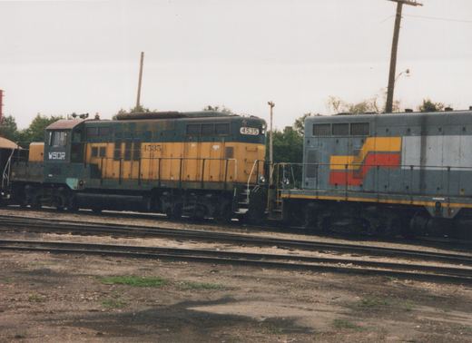 A weathered green and yellow diesel locomotive lettered WSOR and numbered 4535 stands nose to nose with a gray locomotive with red and yellow stripes, on yard tracks under an overcast sky.
