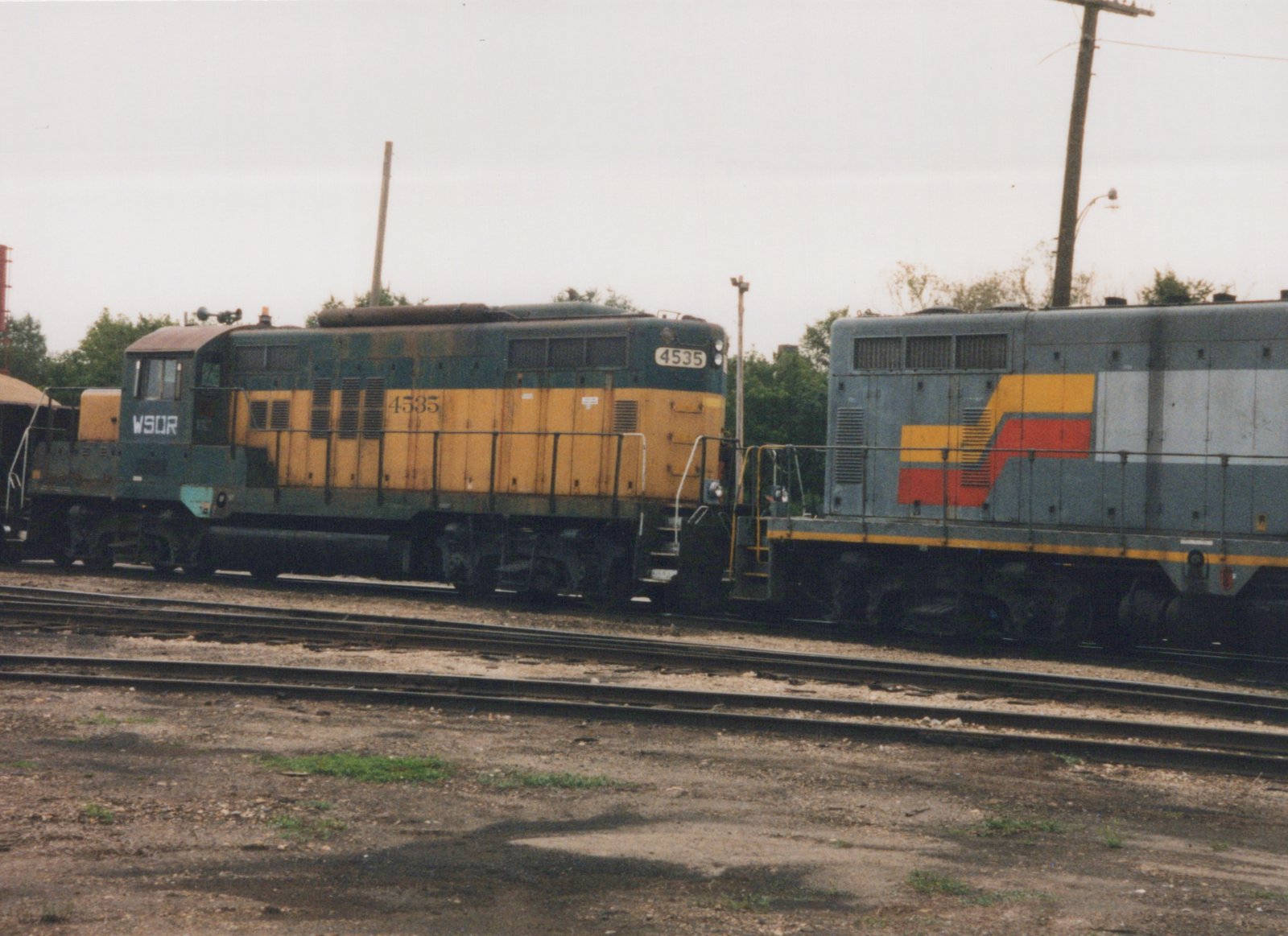 A weathered green and yellow diesel locomotive lettered WSOR and numbered 4535 stands nose to nose with a gray locomotive with red and yellow stripes, on yard tracks under an overcast sky.
