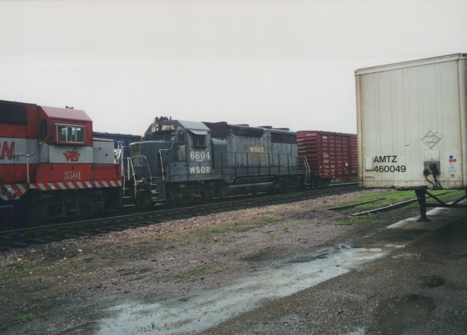A faded blue-gray locomotive lettered WSOR 6604 stands beside the cab of a red and silver locomotive numbered 3501, with a red boxcar behind them and a white highway trailer in the foreground at the right.