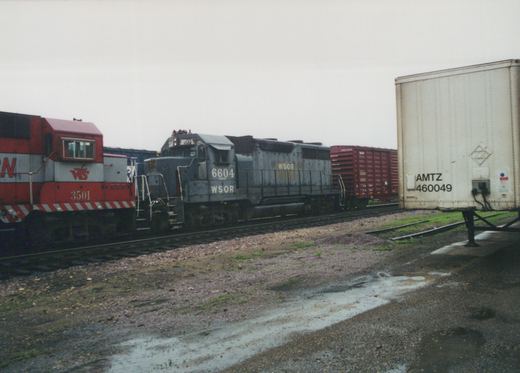 A faded blue-gray locomotive lettered WSOR 6604 stands beside the cab of a red and silver locomotive numbered 3501, with a red boxcar behind them and a white highway trailer in the foreground at the right.