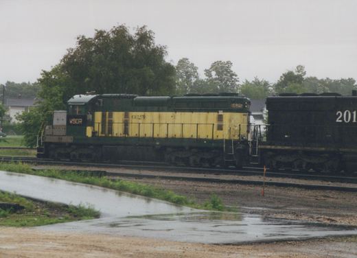 A green and yellow diesel locomotive lettered WSOR, numbered 6625, coupled to a black locomotive on yard tracks under an overcast sky, with wet pavement in the foreground.