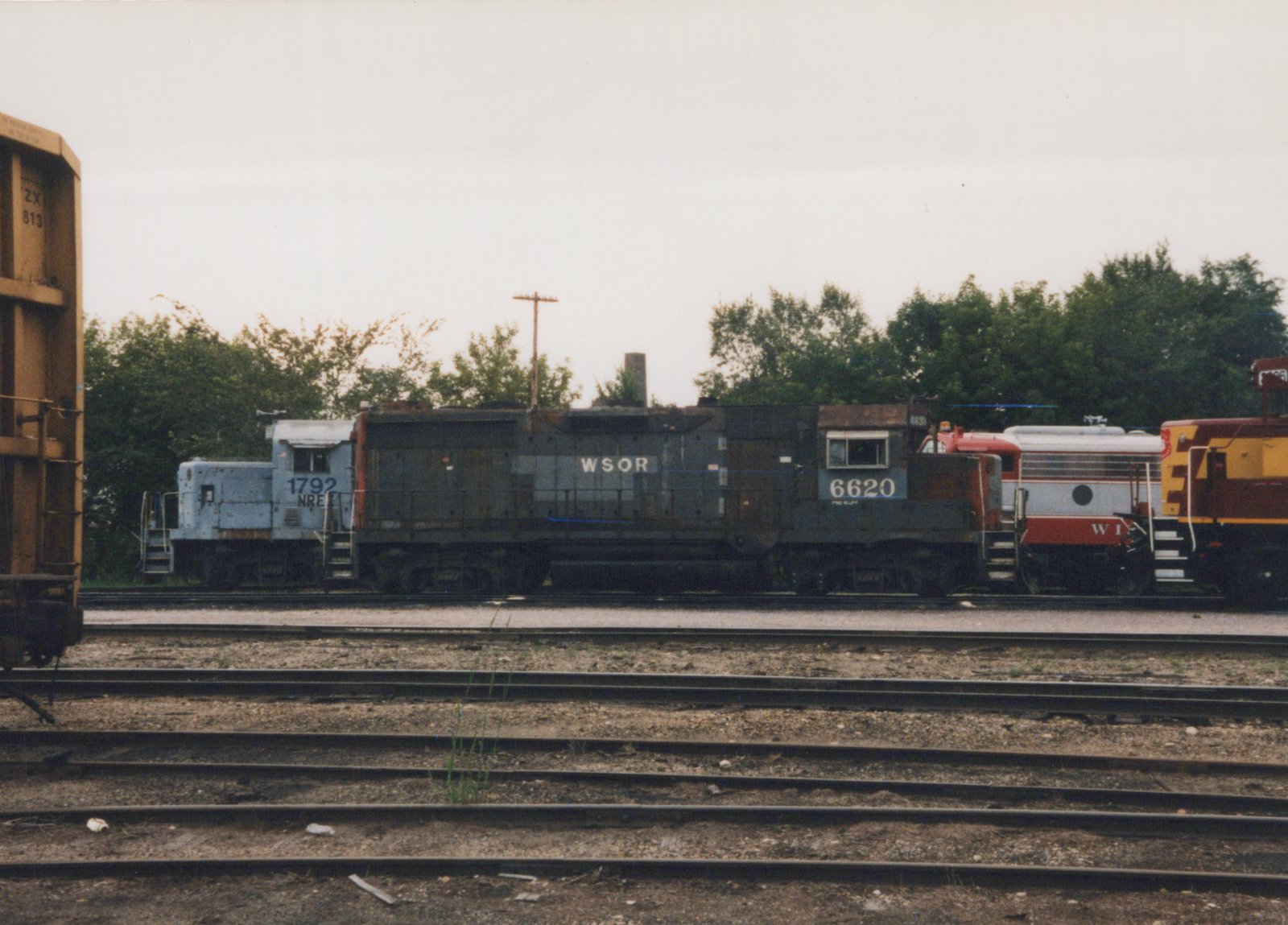 A dark gray diesel locomotive lettered WSOR 6620, side view, coupled between a light gray switcher numbered 1792 and a red and silver cab unit, with a maroon and gold locomotive at the right, seen across empty yard tracks on an overcast day.