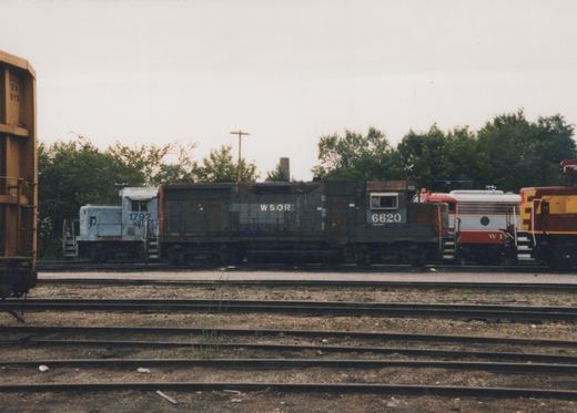 A dark gray diesel locomotive lettered WSOR 6620, side view, coupled between a light gray switcher numbered 1792 and a red and silver cab unit, with a maroon and gold locomotive at the right, seen across empty yard tracks on an overcast day.