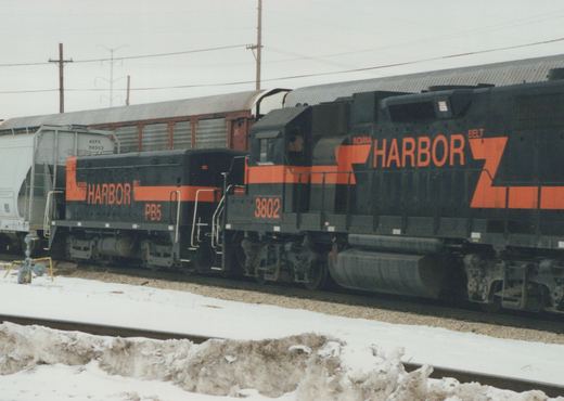 A black and orange Indiana Harbor Belt diesel locomotive, number 3802, coupled to a low cabless unit numbered PB5, in front of autoracks and a gray covered hopper on a snowy, overcast day.