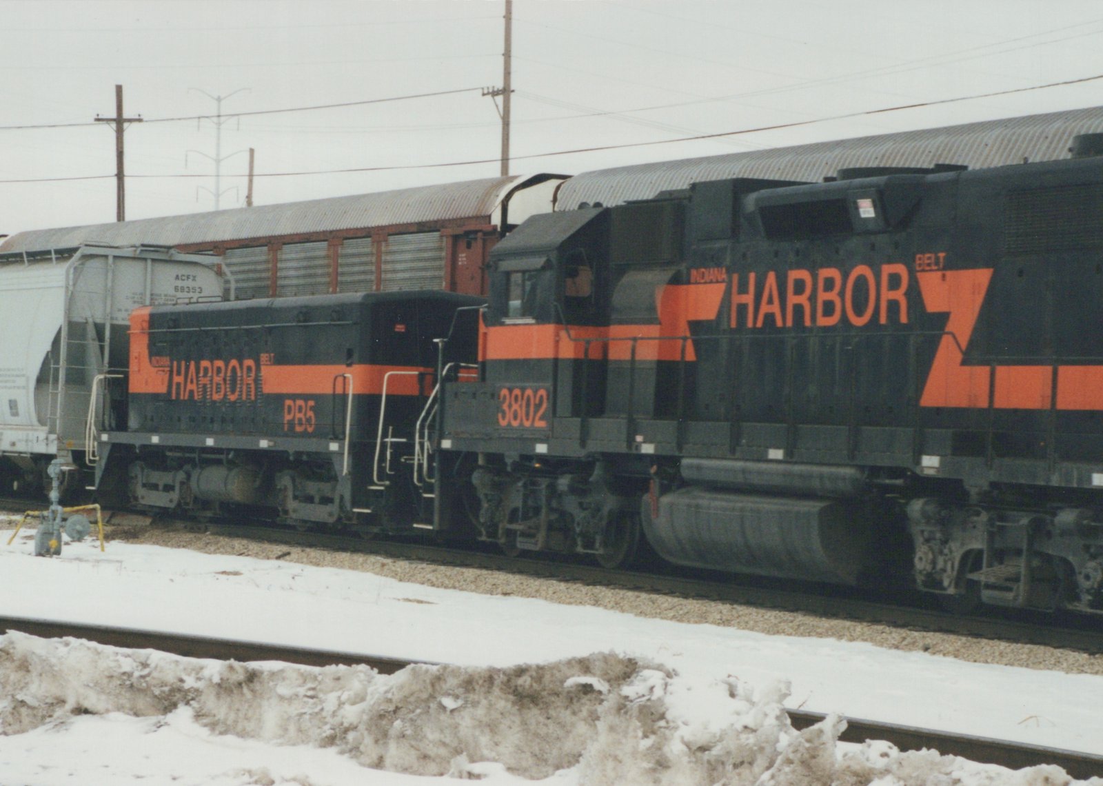 A black and orange Indiana Harbor Belt diesel locomotive, number 3802, coupled to a low cabless unit numbered PB5, in front of autoracks and a gray covered hopper on a snowy, overcast day.