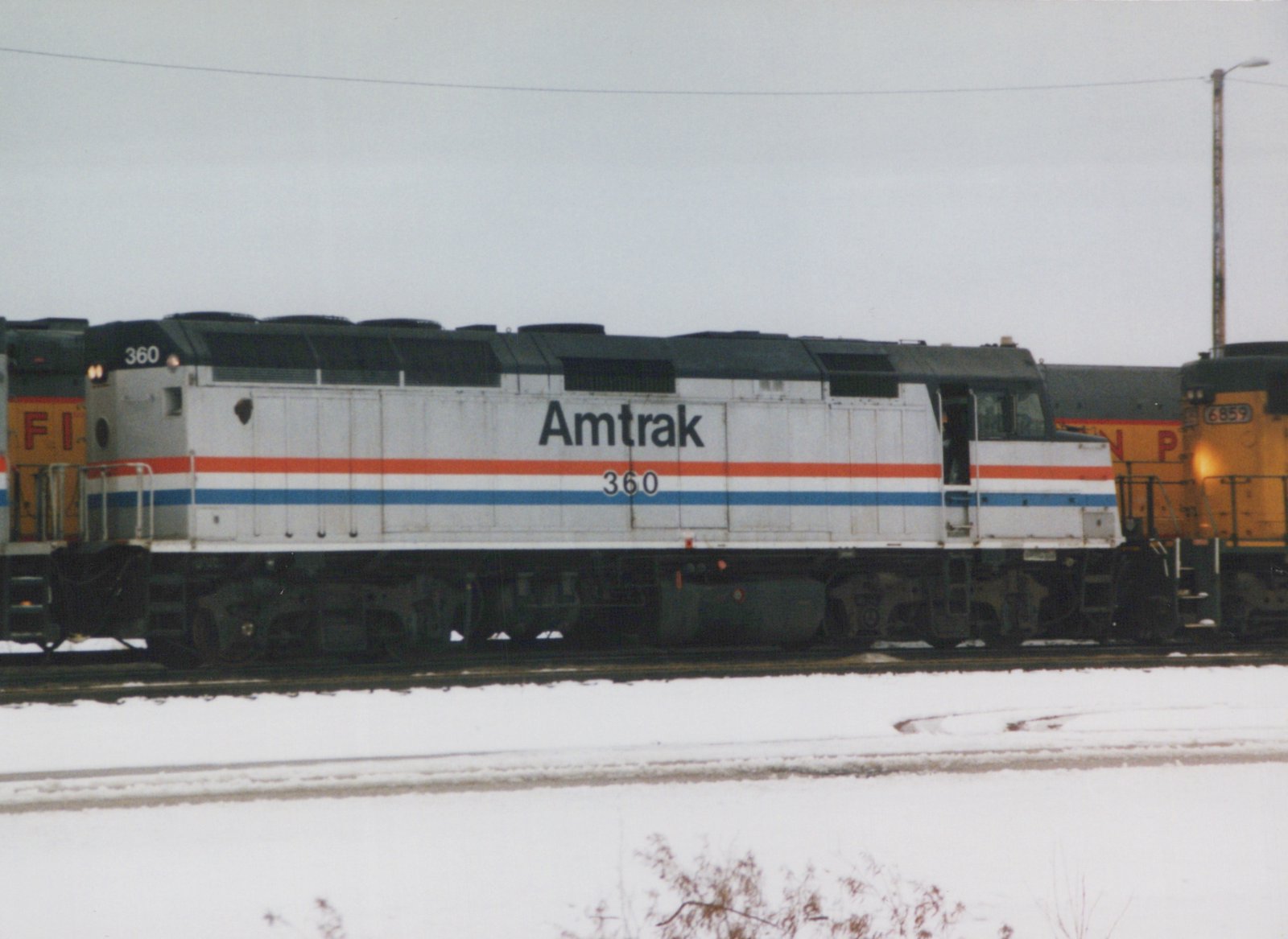 A silver Amtrak diesel locomotive with red, white and blue stripes and the number 360, side view, between yellow locomotives on a snowy yard track under a gray sky.