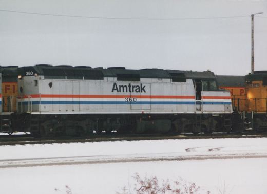A silver Amtrak diesel locomotive with red, white and blue stripes and the number 360, side view, between yellow locomotives on a snowy yard track under a gray sky.