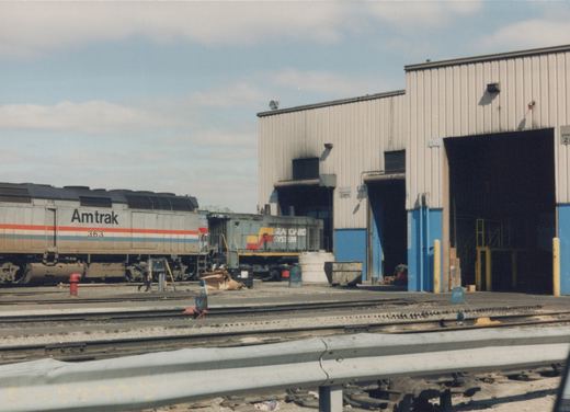 A silver Amtrak diesel locomotive numbered 363 with red, white and blue stripes stands beside a gray Seaboard System switcher in front of a beige metal shop building with blue door frames, seen across several yard tracks and a guardrail.