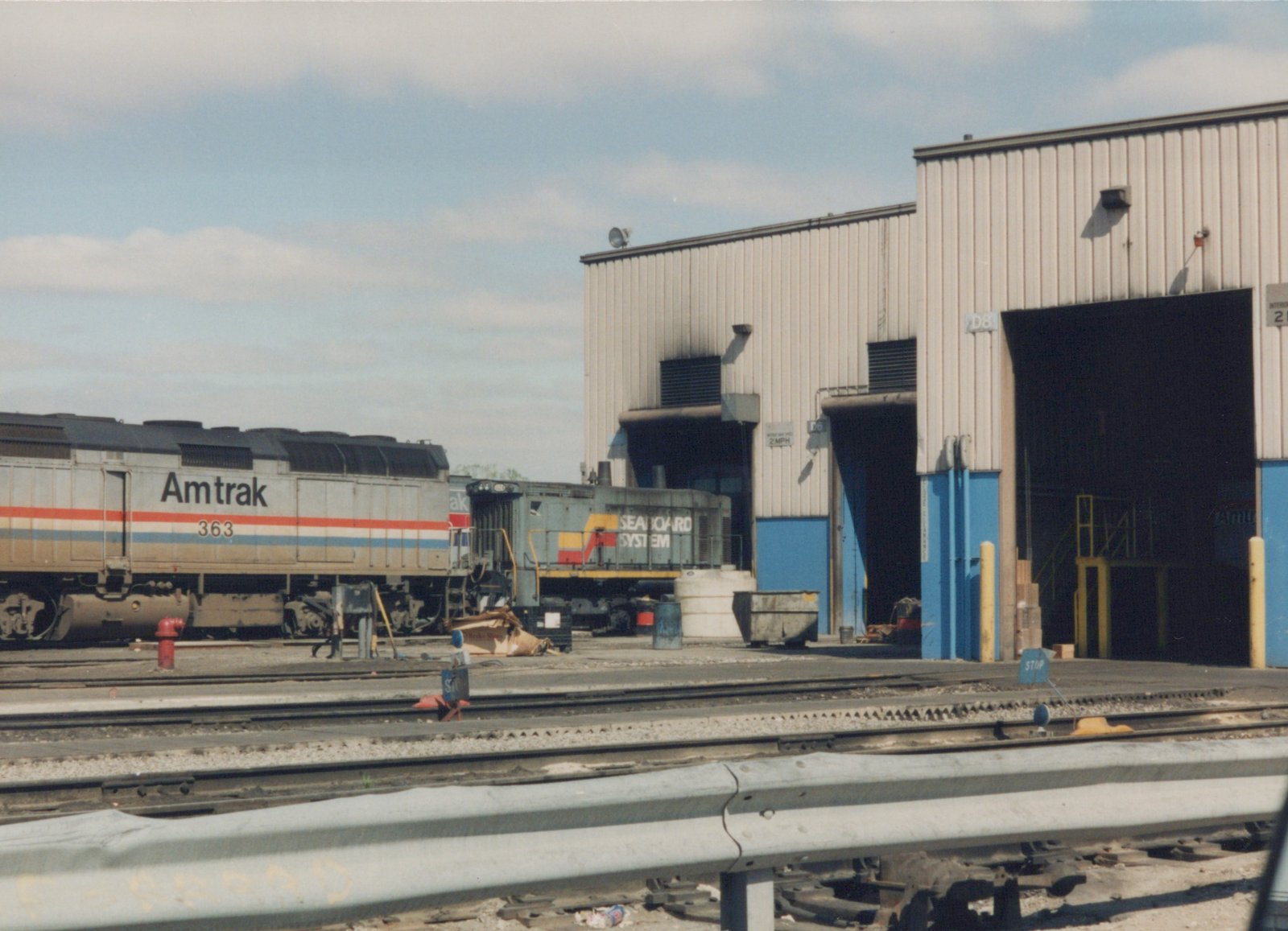 A silver Amtrak diesel locomotive numbered 363 with red, white and blue stripes stands beside a gray Seaboard System switcher in front of a beige metal shop building with blue door frames, seen across several yard tracks and a guardrail.