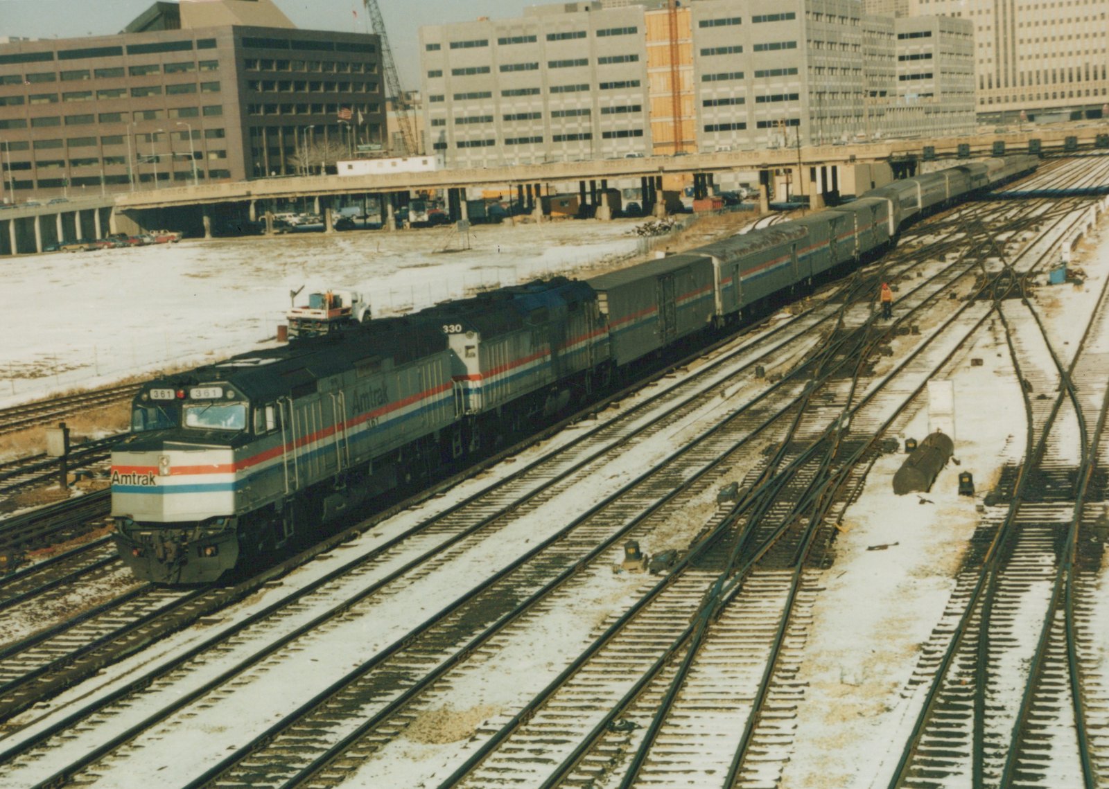 Elevated view of two grey Amtrak diesel locomotives with red, white and blue stripes pulling express cars and passenger cars through a snowy multi-track throat, with a highway viaduct and office buildings behind.