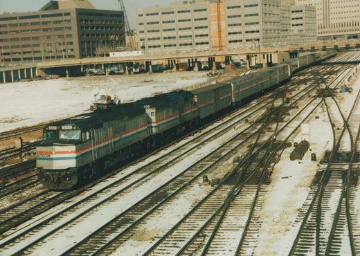 Elevated view of two grey Amtrak diesel locomotives with red, white and blue stripes pulling express cars and passenger cars through a snowy multi-track throat, with a highway viaduct and office buildings behind.
