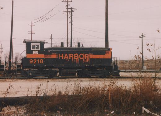 A black diesel switcher with an orange stripe and large orange HARBOR lettering, number 9218, sits on a yard track behind dry weeds, with utility poles and power lines overhead.