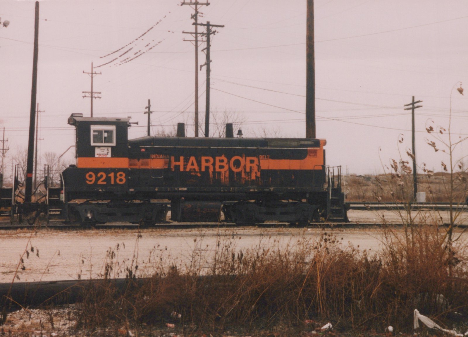 A black diesel switcher with an orange stripe and large orange HARBOR lettering, number 9218, sits on a yard track behind dry weeds, with utility poles and power lines overhead.