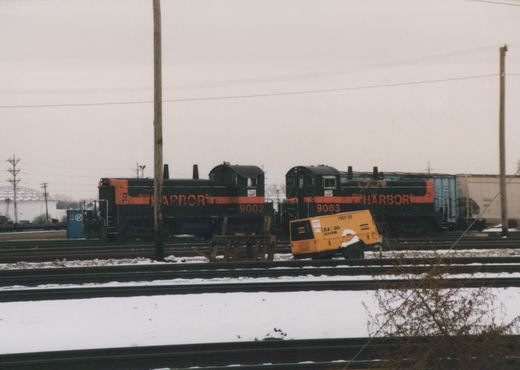 Two black end-cab switcher locomotives with orange stripes and HARBOR lettering, numbered 9002 and 9003, parked cab to cab across several snow-covered yard tracks, with a yellow air compressor in front and freight cars behind.