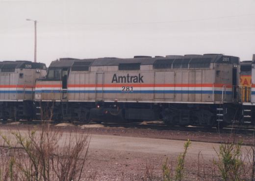 Side view of a silver Amtrak diesel locomotive numbered 281 with red, white and blue stripes, coupled between another Amtrak unit and a yellow locomotive, with brush and gravel in front under an overcast sky.