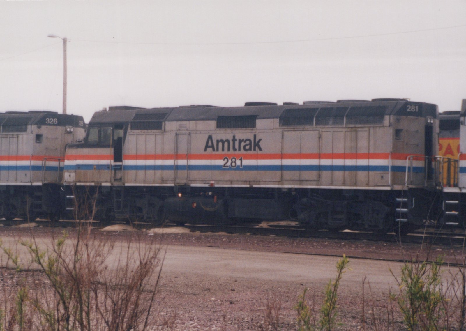Side view of a silver Amtrak diesel locomotive numbered 281 with red, white and blue stripes, coupled between another Amtrak unit and a yellow locomotive, with brush and gravel in front under an overcast sky.