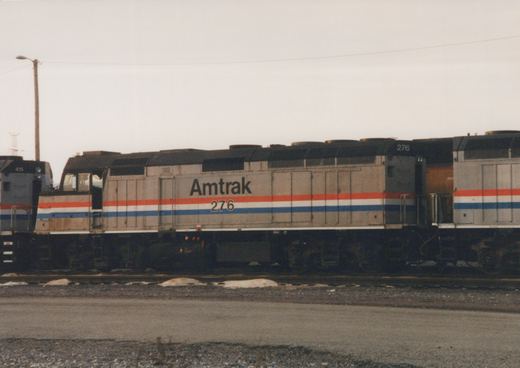 Side view of a silver Amtrak diesel locomotive, number 276, with red, white and blue stripes. It stands between two other locomotives on a yard track, with patches of snow beside the rails under an overcast sky.
