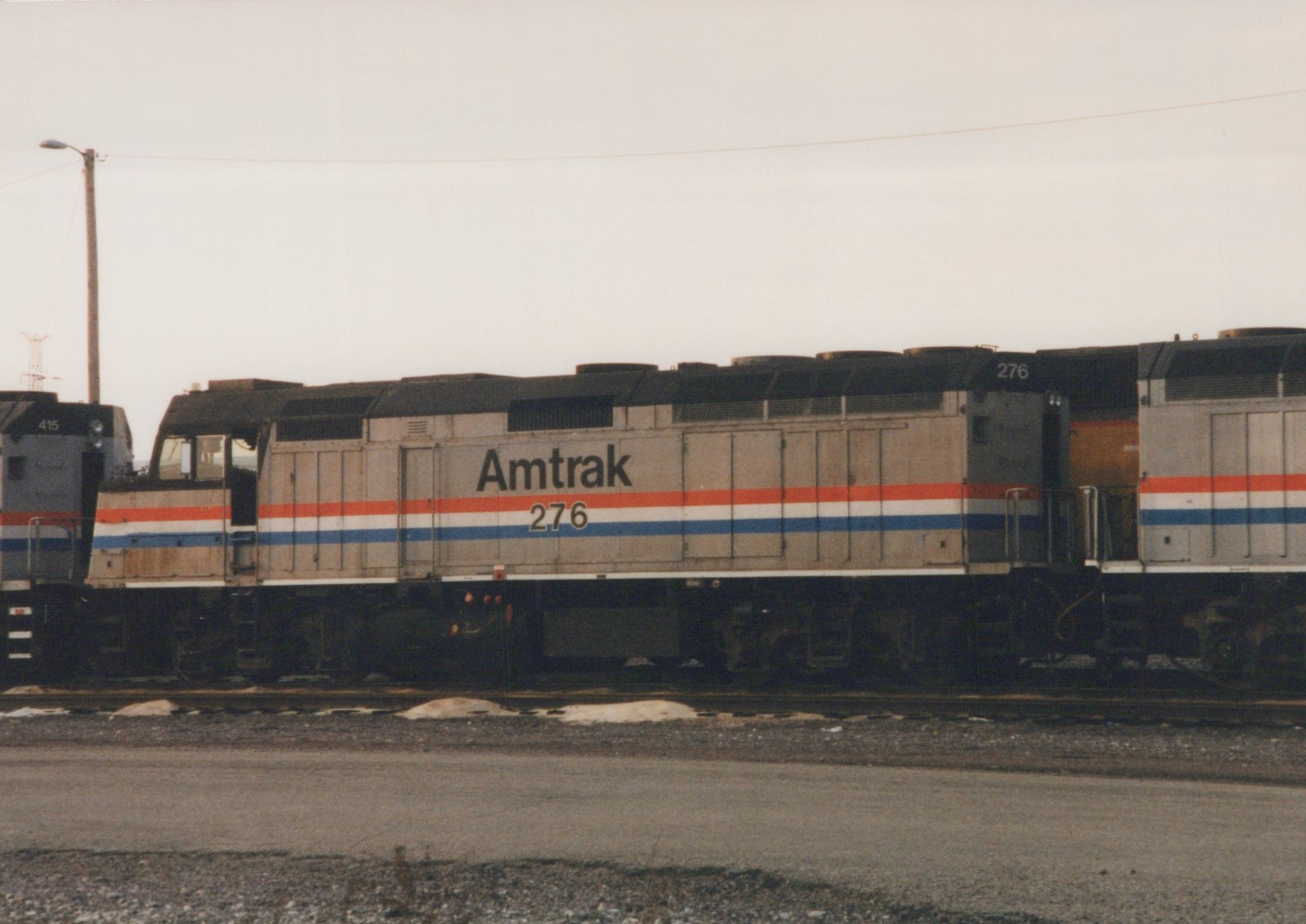 Side view of a silver Amtrak diesel locomotive, number 276, with red, white and blue stripes. It stands between two other locomotives on a yard track, with patches of snow beside the rails under an overcast sky.