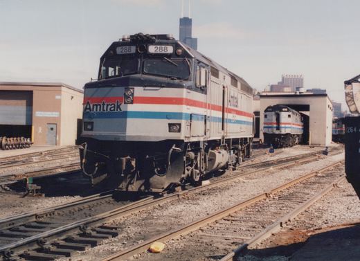 A silver Amtrak diesel locomotive with red, white and blue stripes and number 288 on its number boards stands on yard tracks, with a second Amtrak locomotive numbered 406 inside an open service building and the Sears Tower on the skyline.