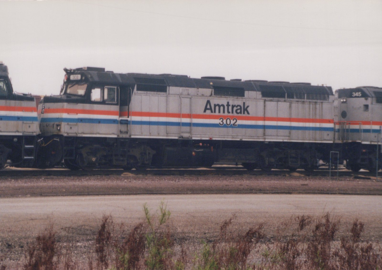 A silver diesel locomotive with red, white and blue stripes, lettered Amtrak 302, stands on a yard track between two similar locomotives under a gray sky, with weeds in the foreground.