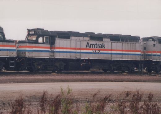 A silver diesel locomotive with red, white and blue stripes, lettered Amtrak 302, stands on a yard track between two similar locomotives under a gray sky, with weeds in the foreground.
