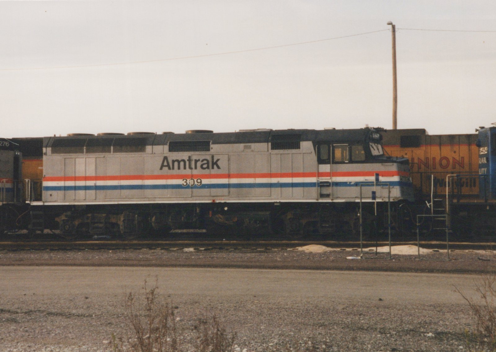 A silver Amtrak diesel locomotive with red, white and blue stripes and number 309 stands sideways in a rail yard, with a yellow Union Pacific locomotive and a blue locomotive behind it, under an overcast sky.